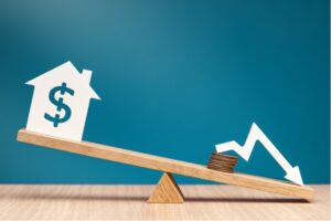 A wooden seesaw balancing a house with a dollar sign against a stack of coins and a falling arrow