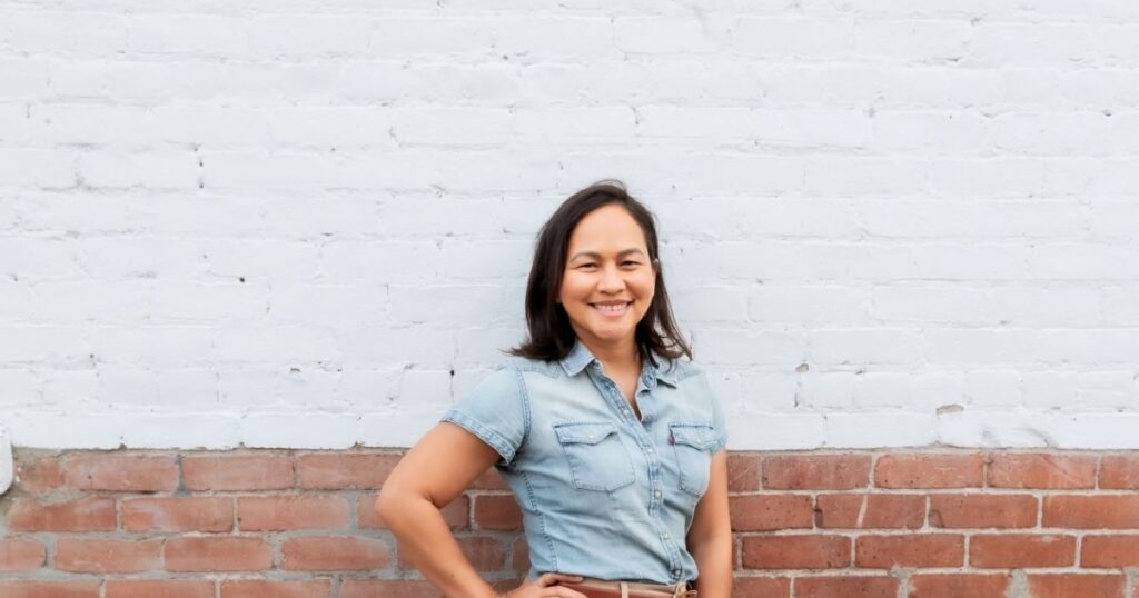 Christine Almarines smiling in a denim shirt in front of a white and red brick wall
