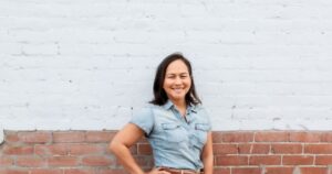 Christine Almarines smiling in a denim shirt in front of a white and red brick wall