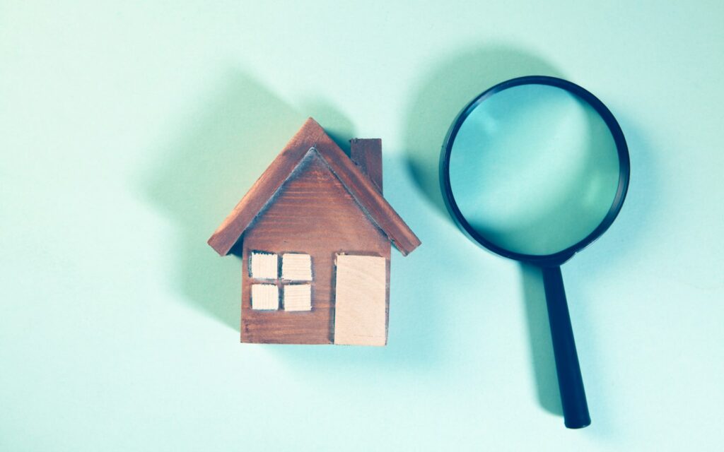 A wooden house model next to a magnifying glass on a teal background