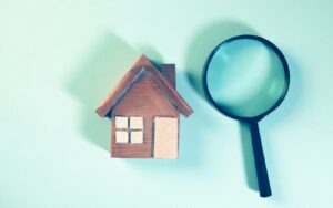 A wooden house model next to a magnifying glass on a teal background