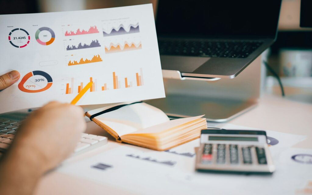 A person pointing a pencil at printed market charts beside a laptop and calculator